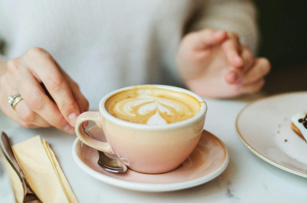 A close-up of a latte with intricate art in a café setting, featuring hands and a saucer.